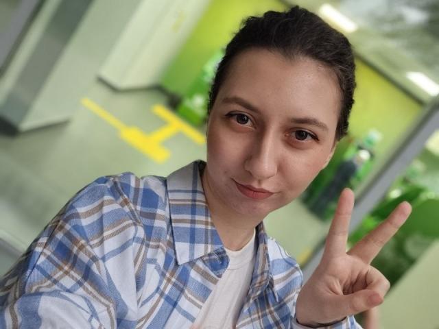 Nichole Kanzenbach, met bruine ogen, donker haar in een knot, gekleed in een blauw geruit shirt over een wit t-shirt, die een vrede-teken maakt, zachtjes lachend, in een kamer met groene muren..