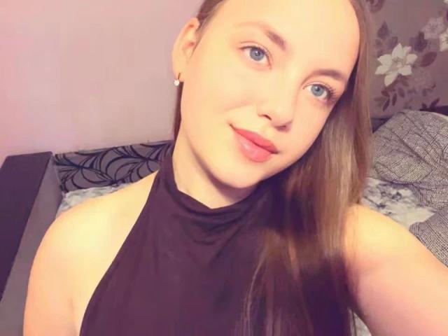 CherryMiss-hot hat blaue Augen, rote Lippen und langes braunes Haar, trägt ein schwarzes Top mit Halter und zeigt ihre nackten Schultern, während sie in die Kamera lächelt..