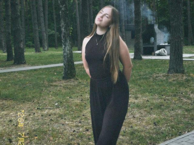 CherryMiss-hot está de pie en un bosque, vestida con un vestido negro sin mangas, con las manos detrás de la espalda, los ojos cerrados, cabello castaño largo, una ligera sonrisa, un collar pequeño, hierba y árboles alrededor..
