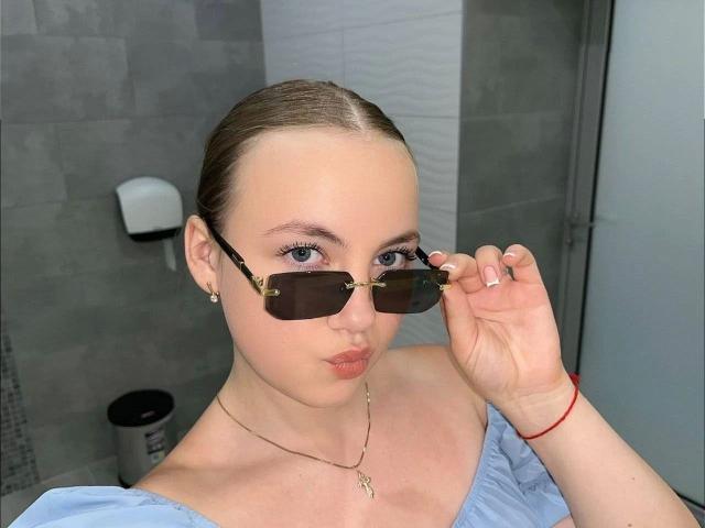 CherryMiss: atractiva, con labios carnosos, ojos azules y cabello rubio recogido en una coleta, sosteniendo gafas de sol negras, vistiendo una blusa azul claro con un hombro descubierto y una pulsera roja..