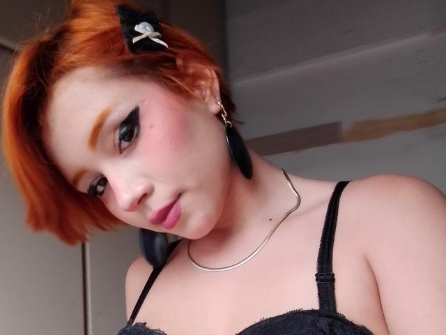 Chloee Williams met oranje haar, een zwarte kanten bh en zwarte oorbel piercings, draagt een zilveren ketting, kijkt naar beneden met een lichte glimlach, heeft roze lippen en dikke zwarte eyeliner..