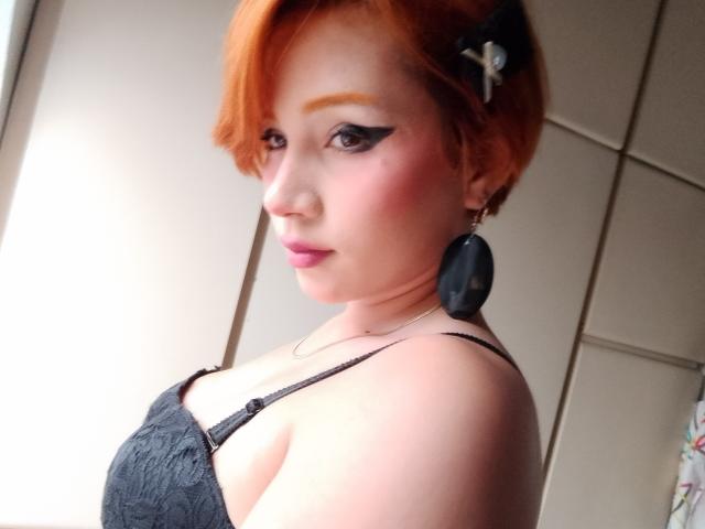 Chloee Williams, con i capelli arancioni, eyeliner nero, fard rosa, vestito nero, orecchini rotondi, che guarda di lato, mostrando il seno, luce soffusa, leggero sorriso, molletta nera tra i capelli..