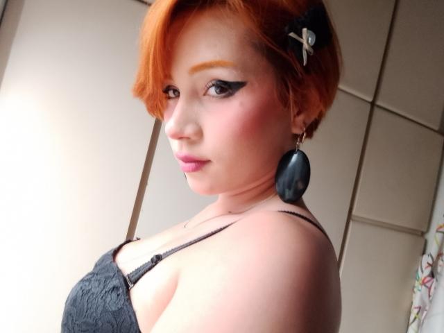 ChloeeWilliams has orange hair, black eyeliner, pink blush, black earrings, and a black lace top, showing her tits and a confident, slightly smoky expression.