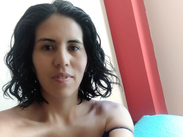 LarisaBrown has short, wavy black hair, light brown skin, and is looking at the camera with a slight smile, showing her bare shoulders and a strap on her right.