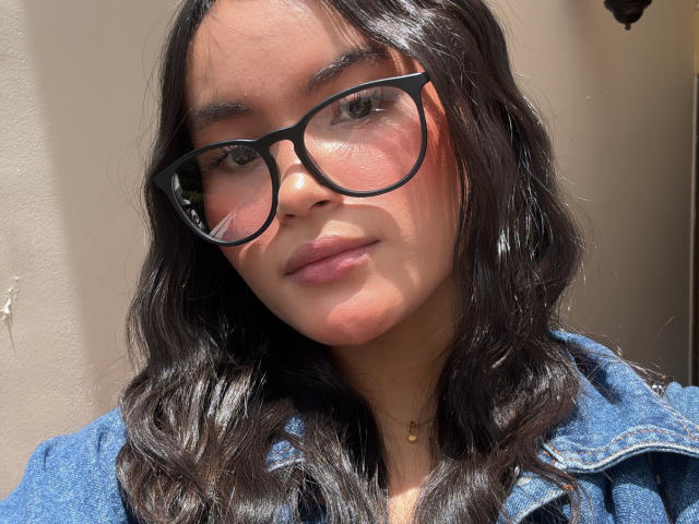 MiiaPalmer with dark wavy hair, wearing black glasses and a blue denim jacket, has a cute face, pink blush, and a small gold necklace, looking slightly to the side.