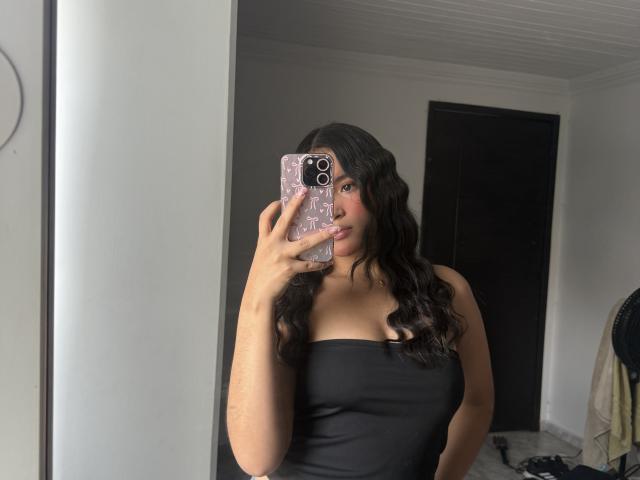 MiiaPalmer with wavy black hair, wearing a black strapless top, holding a pink phone with white patterns, taking a mirror selfie showing her tits and slightly smiling.