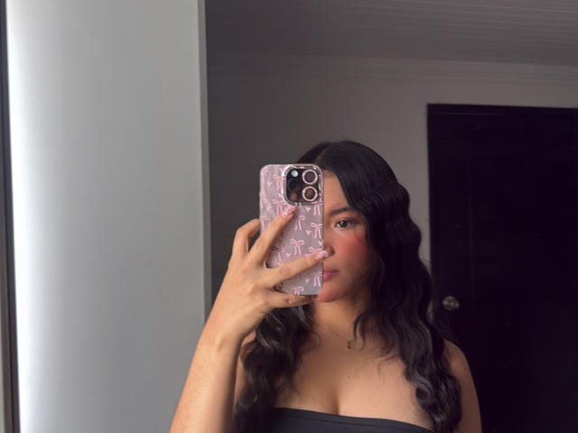 MiiaPalmer with black wavy hair, wearing a strapless black top, holding a pink phone with a textured case, taking a mirror selfie showing her Tits and red lipstick.