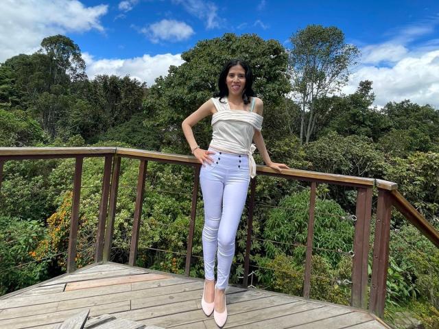 LarisaBrown stands on a wooden bridge, wearing a white off-shoulder top and tight white jeans, with pink heels, her small tits and slim ass visible, smiling.