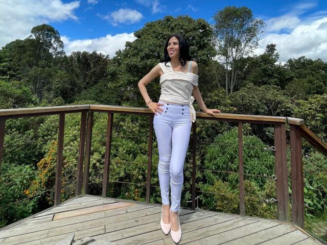 LarisaBrown stands on a wooden bridge, arms resting on the railing, wearing a white off-shoulder top, tight white jeans, and pink heels, with a blue sky and trees.