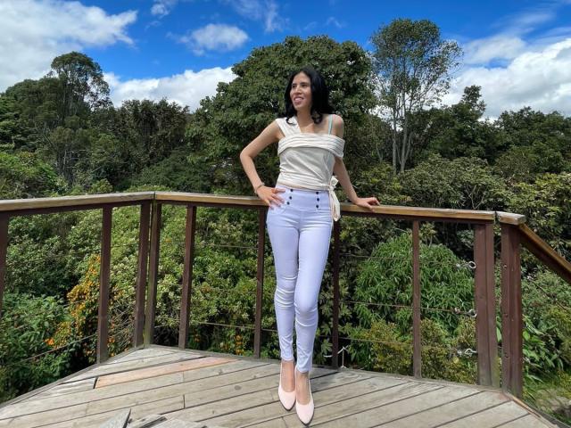 LarisaBrown stands on a wooden deck, hands on railing, wearing a white off-shoulder top, tight light blue jeans, and pink heels, with green forest and blue sky behind.
