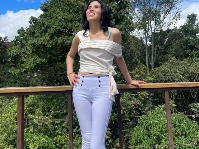 LarisaBrown with black hair, smiling, wearing a white off-shoulder top and white high-waisted pants, standing on a wooden balcony, hands on railing, looking up, butt visible.