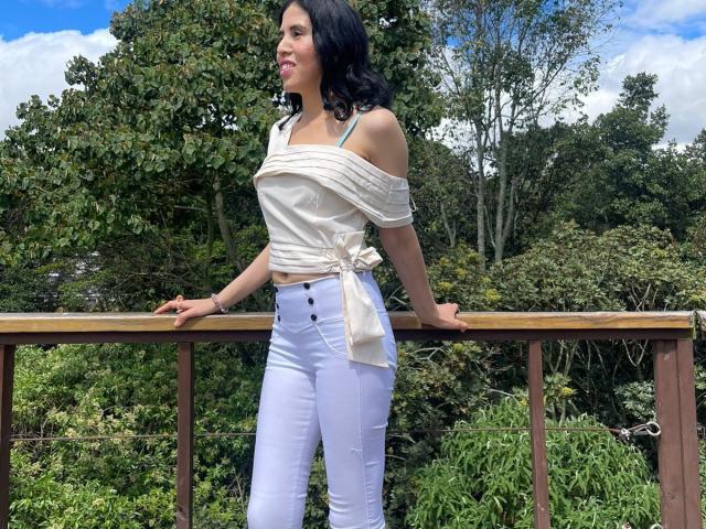 LarisaBrown with black wavy hair, white off-shoulder top, tied at waist, white high-waisted pants, standing on wooden balcony, hands on railing, looking to side, blue sky.