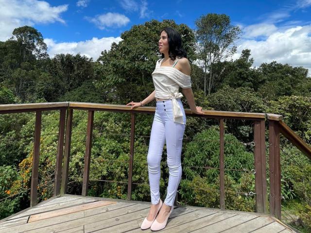 LarisaBrown with small tits, white off-shoulder top, tied at waist, white jeans, pink heels, standing on wooden bridge, arms on railing, black wavy hair, blue sky, green trees.