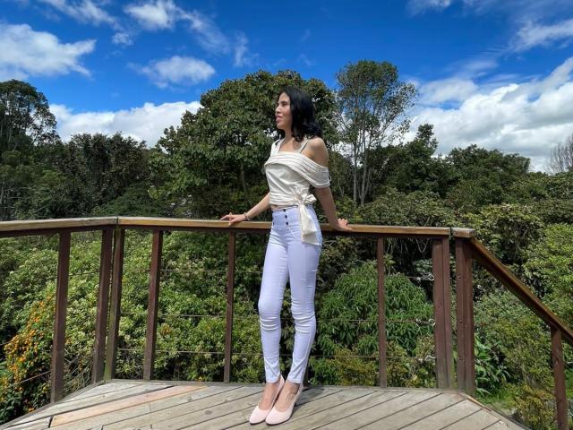 LarisaBrown with white off-shoulder top, tied at the waist, white skinny jeans, and pink heels, standing on wooden bridge, arms on railing, under blue sky with clouds.