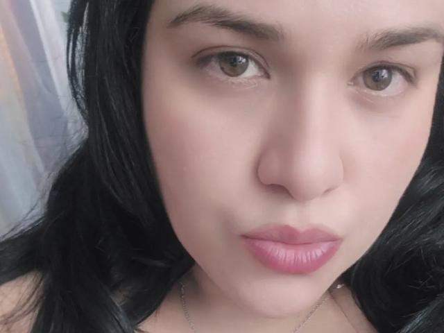 VivianaThomson's face, with pink lipstick, black wavy hair, and brown eyes, looking straight at the camera. She has a nose ring and a silver necklace.