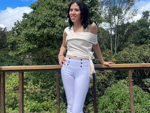 LarisaBrown with black hair, white top tied at waist, off-shoulder, white high-waisted jeans, standing against wooden railing, hands on rail, smiling, green forest background.