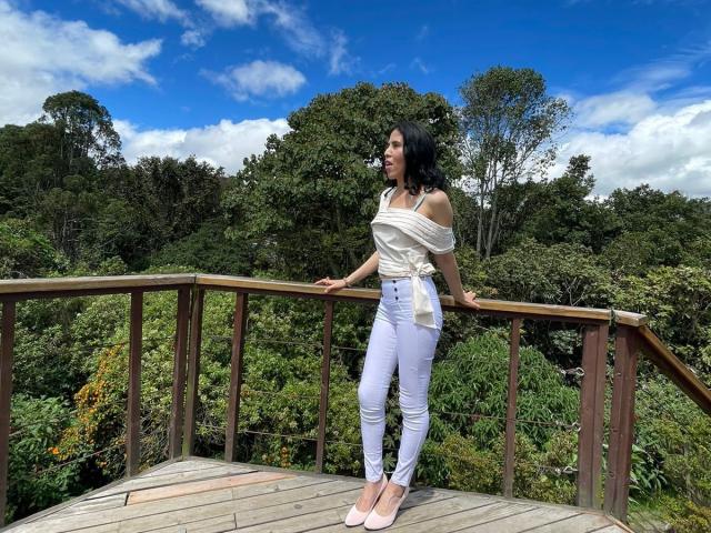 LarisaBrown with black hair, white off-shoulder top, and tight white jeans, standing on wooden bridge, leaning on railing, wearing pink heels, against lush green forest.