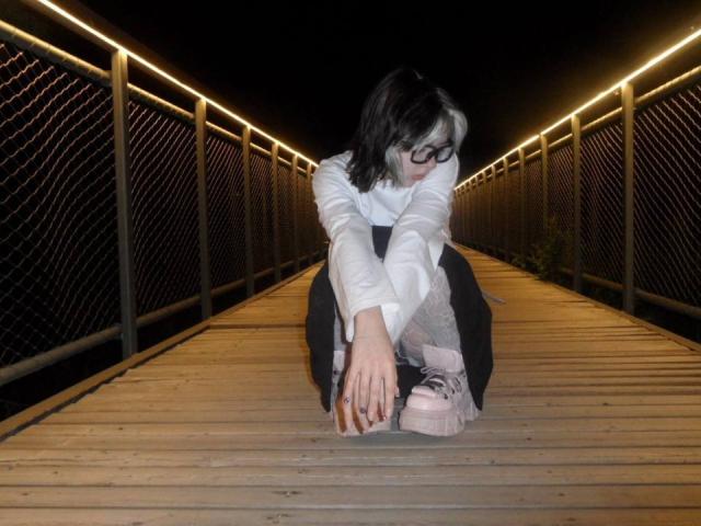 KrystalBerkovitch is squatting on a wooden bridge at night, stretching her hands towards her pink sneakers, wearing a white long-sleeve shirt and black pants.