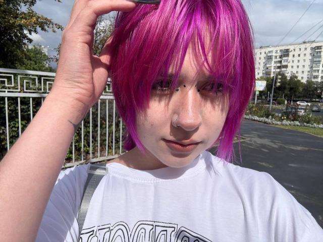 KrystalBerkovitch with bright purple hair, freckles, and a nose ring, wearing a white shirt, holding her hair up, standing outdoors with a serious expression.