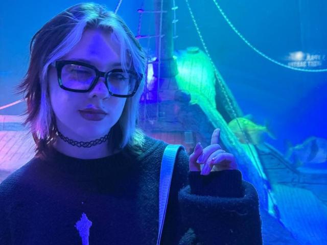 KrystalBerkovitch with blue-tinged hair, black glasses, dark lipstick, black choker, black sweater, and finger in the air, looking confident with a slight smile. Her tits are small.