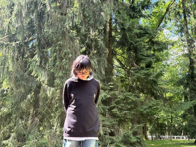 KrystalBerkovitch stands in a forest, wearing a black long-sleeve shirt and blue jeans, with hands in pockets, and has dark brown hair and glasses.