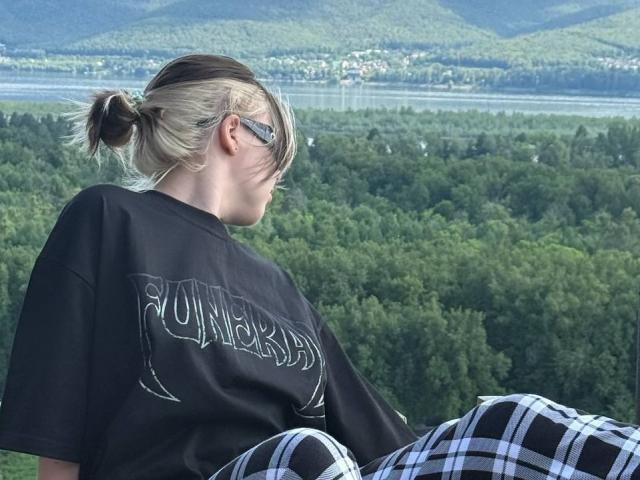 KrystalBerkovitch with blonde hair in a ponytail, wearing a black t-shirt and black-and-white plaid pants, sitting and looking away. She has glasses and a small, white earpiece.