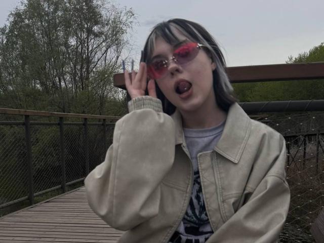 KrystalBerkovitch with pink sunglasses, tongue out, wearing a beige jacket over a gray tee, and gray-striped hair, touching her face on a wooden bridge.