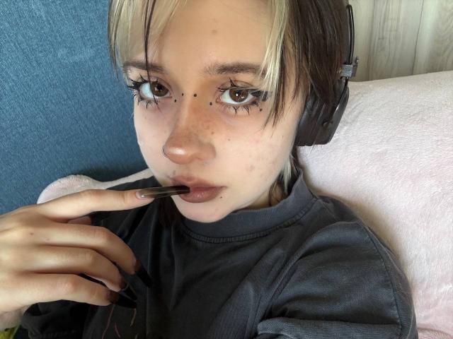 KrystalBerkovitch with blonde and brown hair, wearing headphones, eating a chocolate chip, has black eyeliner and small freckles, looking at camera, wearing a gray shirt, on pink chair.