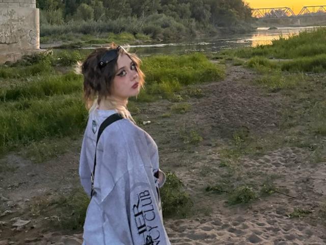 KrystalBerkovitch stands on a sandy path, wearing a loose white shirt, with sunglasses on her head, looking over her shoulder, showing her tits, in a grassy, river-side setting.