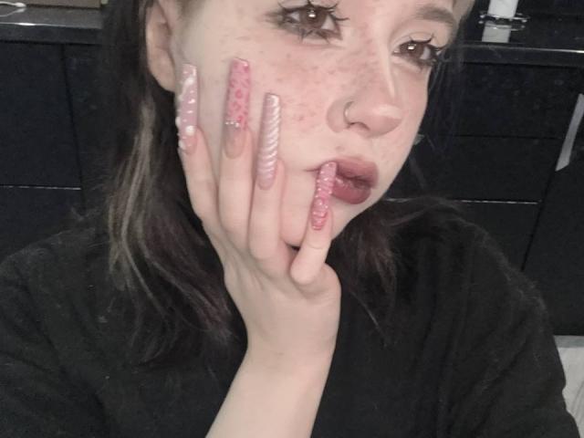 KrystalBerkovitch with pink and white striped acrylic nails, freckled face, and dark lipstick, sucking her fingers, wearing a black shirt, in a dimly lit room.
