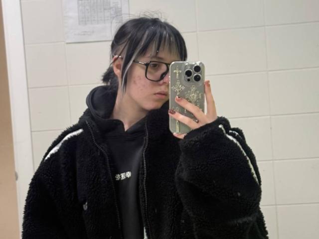 KrystalBerkovitch with black hair, glasses, and a black fuzzy jacket, taking a selfie in a bathroom, holding a phone with a green case, showing a pouty face.