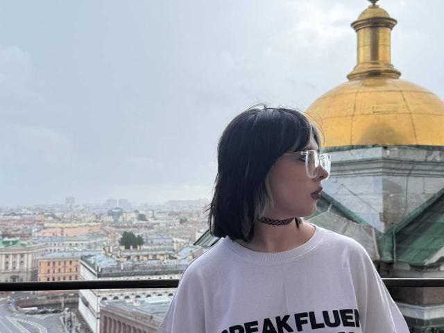 KrystalBerkovitch with black hair, glasses, white tee, black choker, and "peak fluen" text, standing by a golden dome, looking to the right, cityscape in the background.