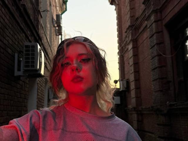 KrystalBerkovitch with wavy blonde hair, wearing a gray sweater, lit by red and blue light, standing in a narrow brick alleyway at sunset.