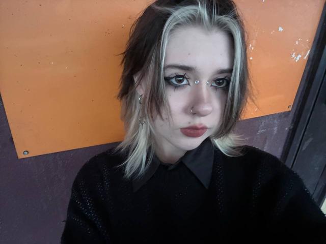 KrystalBerkovitch has pale skin, blonde and brown hair, black eyeliner, nose ring, and is wearing a black sweater with a collared shirt. She's looking down with pouty red lips.