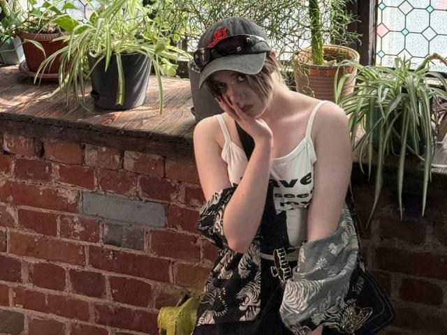 KrystalBerkovitch with pale skin, short brown hair, white tank top, black jacket, black pants, baseball cap, hand on cheek, sitting against red brick wall with potted plants.