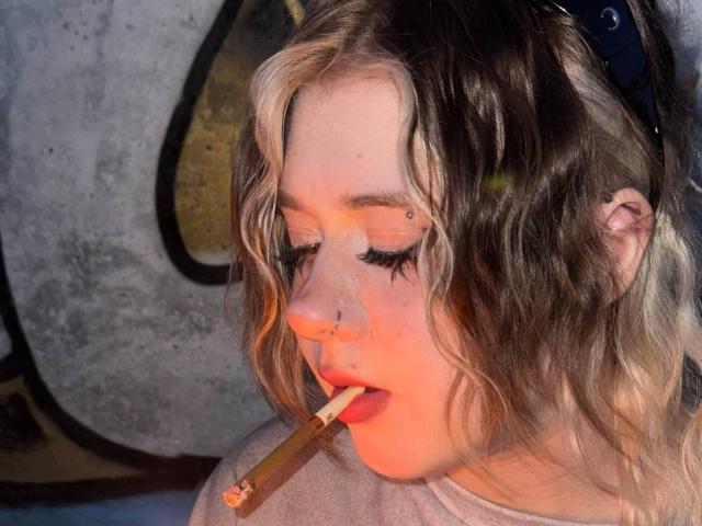 KrystalBerkovitch with wavy blonde and brown hair, nose ring, and freckles, holding a cigarette in her slightly open mouth, eyes closed, wearing a grey shirt.