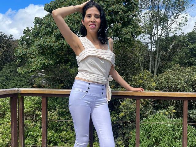 LarisaBrown with dark hair, smiling, wearing a white strapless top and tight white pants, standing on a wooden balcony, hand in hair, blue sky, green trees in background.