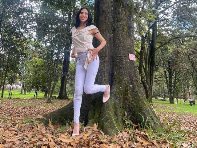 LarisaBrown standing with one leg up against a tree, wearing a white top, tight light purple jeans, and pink heels. Her black hair is down, smiling. Tree in foreground.