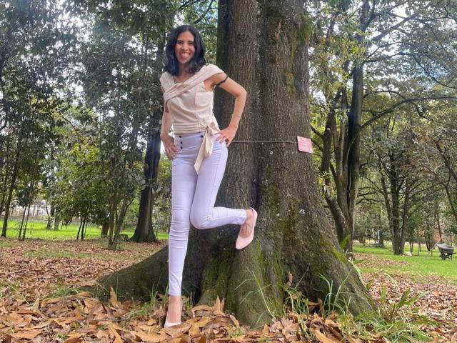 LarisaBrown stands with one leg up, leaning on a tree, wearing a beige top, white pants, pink flats, smiling. Tree roots, fallen leaves, and forest in background.
