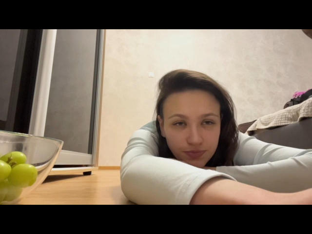 IsobelStaehle ligt op een houten tafel, met haar armen over elkaar, en draagt een lichtblauwe blouse. Aan de linkerkant staat een glazen schaal met groene druiven. Ze heeft bruin haar en een neutrale uitdrukking..