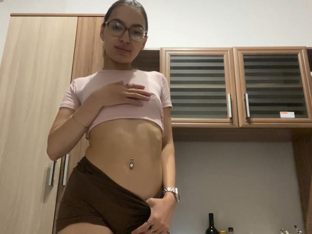 RaneeDerienzo with glasses, pink crop top, brown shorts, flat stomach, navel piercing, and hand on chest, standing in kitchen, looking at camera.