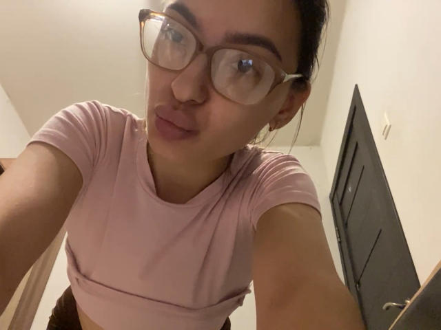 RaneeDerienzo with glasses, pink t-shirt, and pouty lips, looking down at the camera, showing her small tits, brown skin, and dark hair in a messy bun.