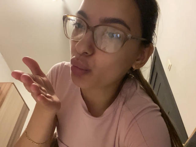 RaneeDerienzo, with glasses and pouty lips, puffs out her right cheek, wearing a pink shirt, brown hair in a ponytail, small hoop earrings, in a dimly lit room.