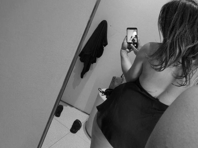 Selfie em preto e branco de Miky69I, com as costas viradas para a câmera, usando uma saia, mostrando o bumbum nu, segurando o celular na mão direita, cabelo comprido, casaco na parede, chinelos no chão..
