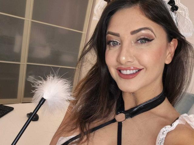 Miky69I mit dunkelbraunen Haaren, weißem Spitzen-Haarreif für Dienstmädchen, schwarzem Top mit Halter, weißem Staubwedel, lächelnd in die Kamera, trägt rosa Lippenstift und schwarzen Augen-Make-up, in einem Zimmer..
