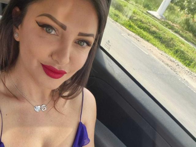 Miky69I con rossetto rosso, vestito viola, eyeliner ad ali di farfalla e collana, seduta in macchina, che guarda la telecamera, mostrando il seno e un sorriso carino..