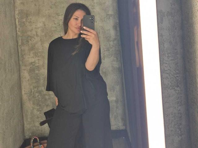 Miky69I hat langes, braunes Haar, ein süßes Gesicht und trägt ein lockeres schwarzes Hemd und eine Hose. Sie macht ein Selfie vor einem Spiegel mit einem Smartphone in einem grauen Raum..