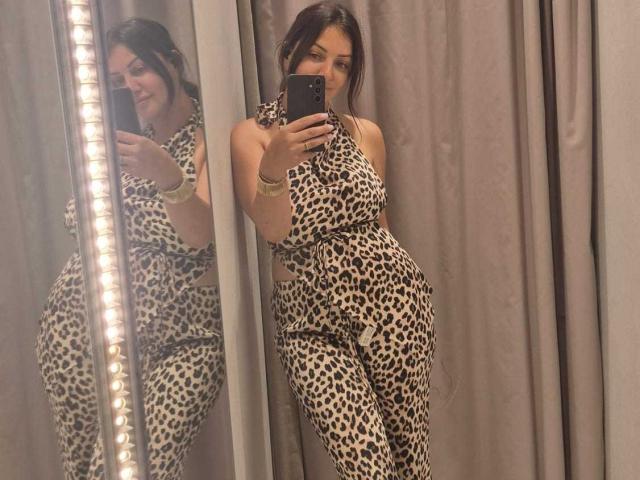 Miky69I con un vestito a stampa leopardo, che tiene un telefono, mostrando il riflesso nello specchio, sorridendo, formosa, con un seno di media grandezza, capelli castani, tenda grigia, che indossa un braccialetto dorato, in piedi, luci dello specchio..