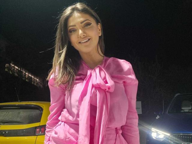 Miky69I, con lunghi capelli castani, sorridente, indossa un abito rosa con un fiocco, in piedi di fronte a auto gialle e blu di notte. Il seno è leggermente visibile sotto l'abito..