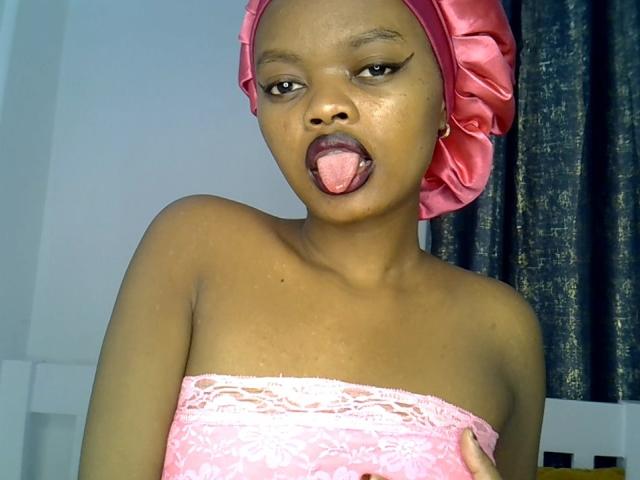 NaughtyPatricia with dark skin, pink lace strapless top, pink headscarf, tongue out, showing her bare shoulders, and a slight hand on her tit. Her expression is playful and seductive.